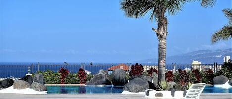 A pool area with a view of the ocean, palm trees, and a fence.