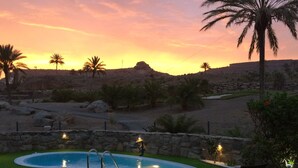 Une piscine bordée d’un mur de pierre, une pelouse et un coucher de soleil en arrière-plan.