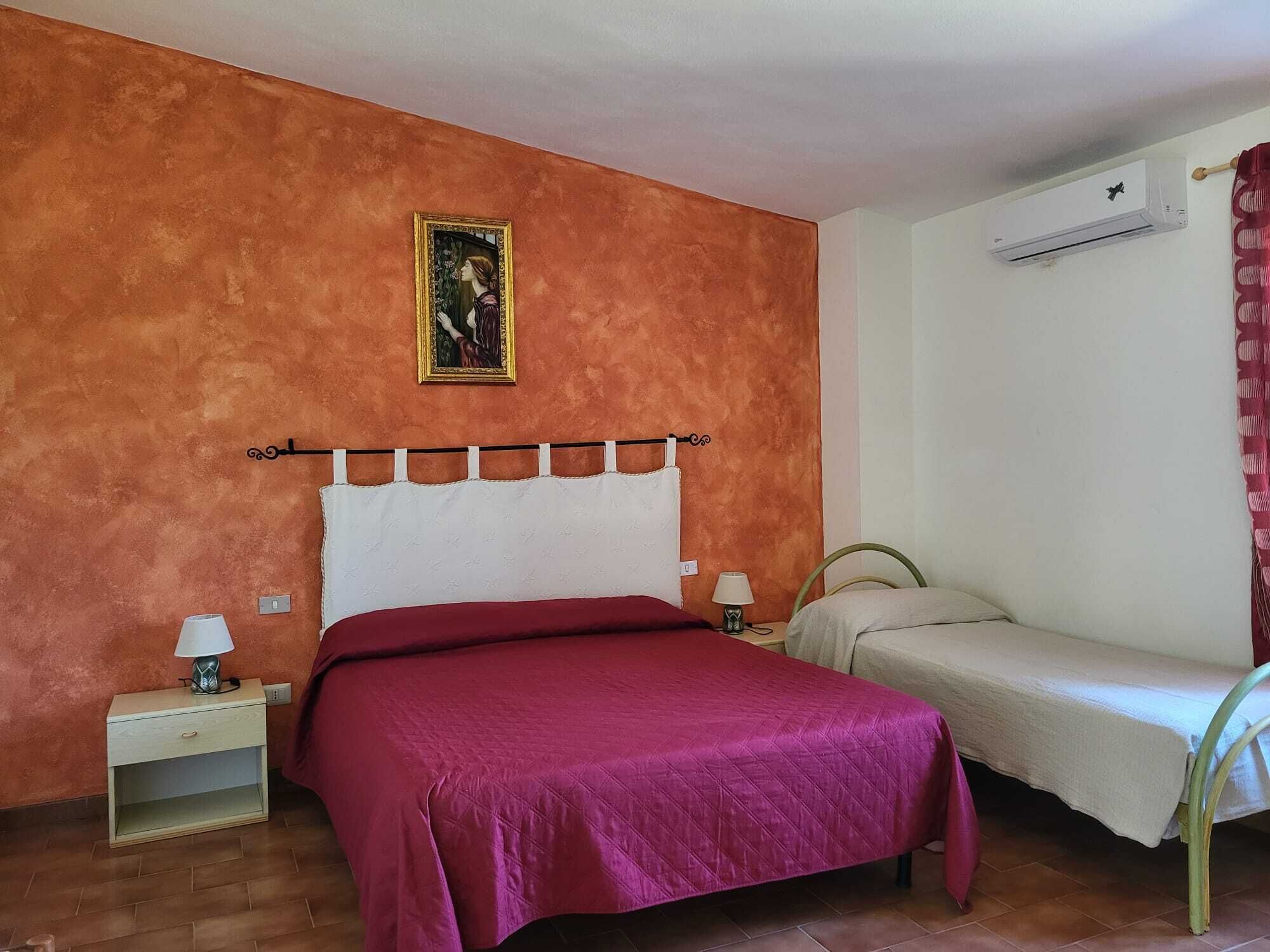 Ein Schlafzimmer mit einem großen Bett, einem kleineren Bett, einer Klimaanlage, einem Nachttisch mit Lampe und einem Bild an der Wand.
