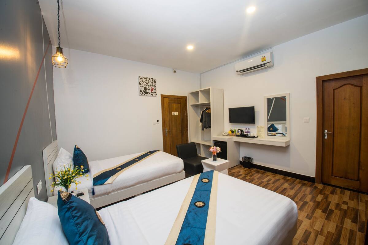 Habitación de hotel con dos camas, un escritorio, una silla, una mesita con un florero y un espejo, además de un equipo de aire acondicionado.