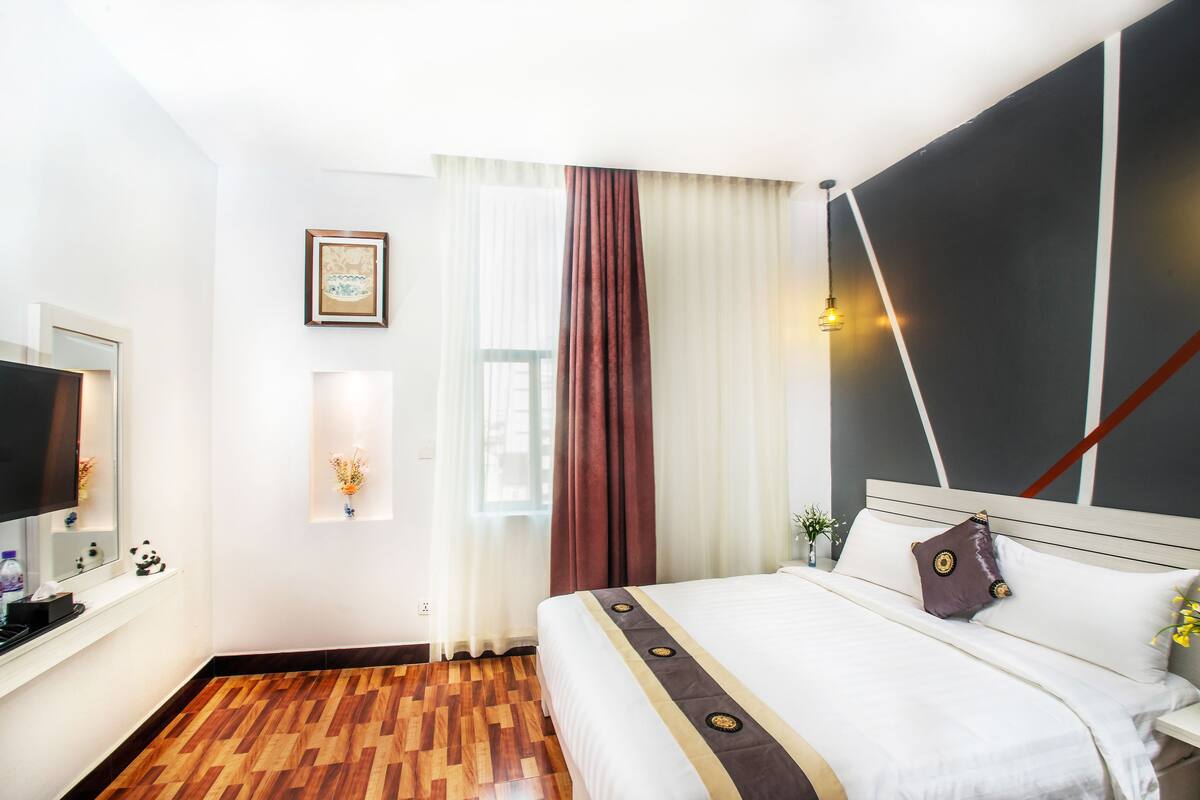 Habitación de hotel con piso de madera, cama con ropa blanca y almohadas decorativas, mesita de noche con lámpara, ventana con cortinas translúcidas y televisor montado en la pared.