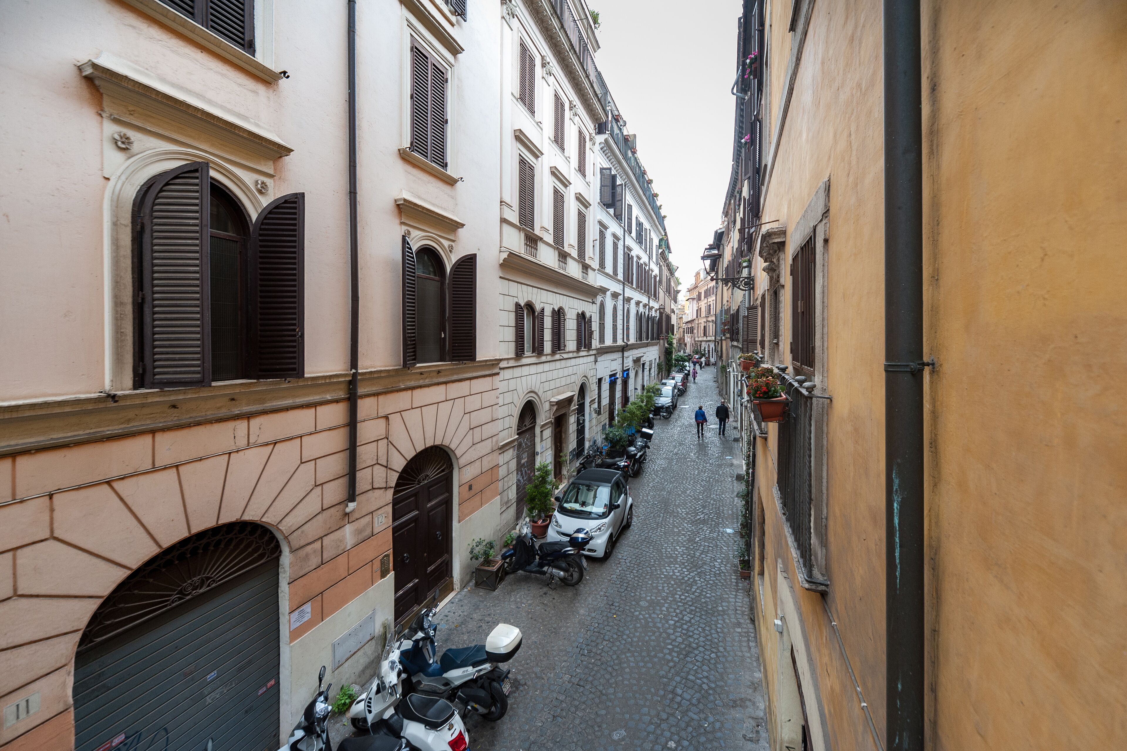 A narrow cobblestone street lined with historic buildings, parked scooters, and a few pedestrians.