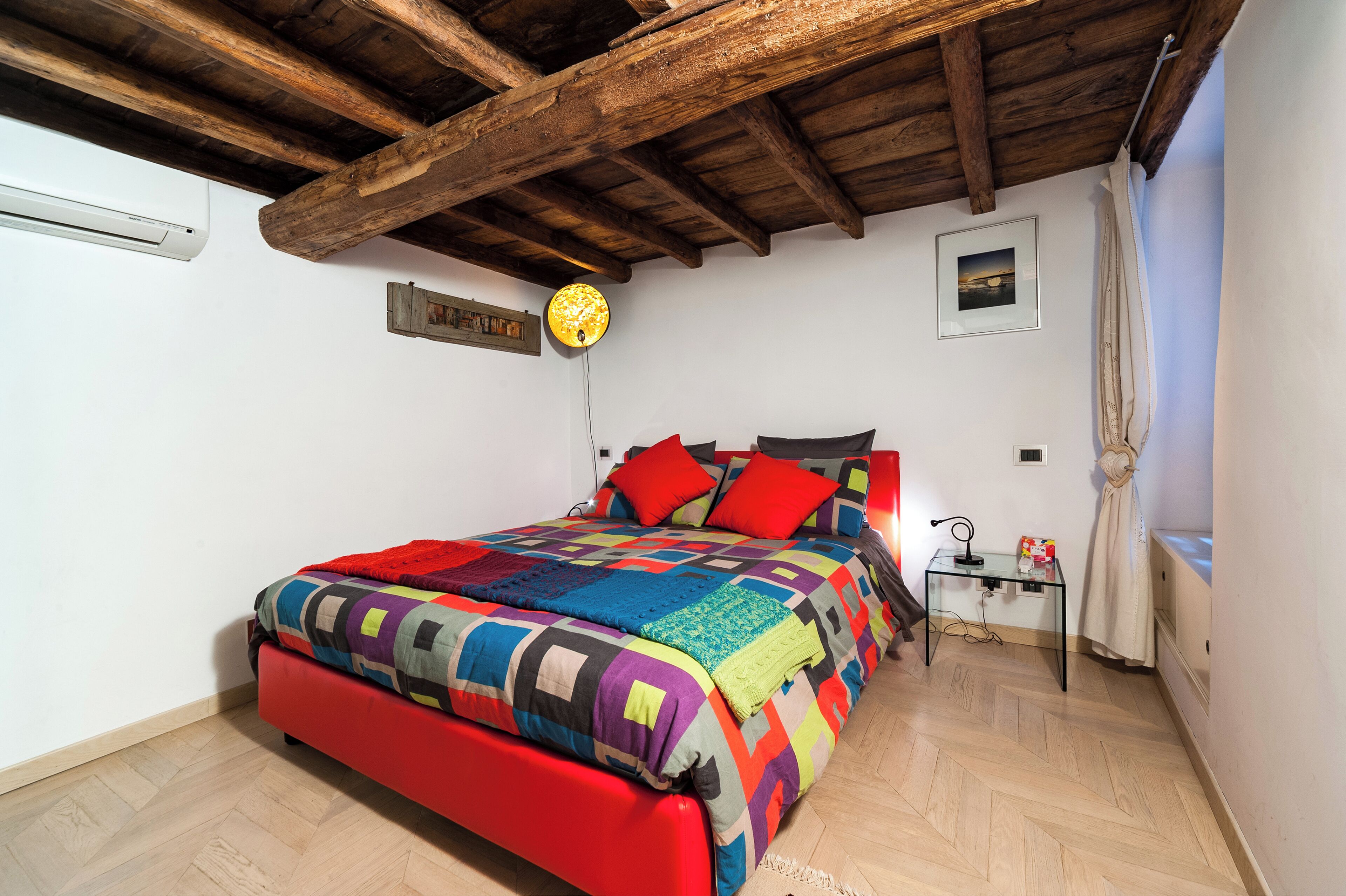 A bedroom with a wooden ceiling, a colorful quilted bedspread, a red bed frame, a small bedside table with a lamp, and a framed picture on the wall.