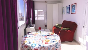 A room with a dining table covered with a tablecloth featuring kitchen utensils, a red sofa, a window with purple curtains, and a door with framed pictures. - Apartment with terrace, sea views, and satellite TV via Wi-Fi 600 MB (Gran Tarajal)