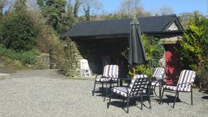 Een grind terras met vier gestreepte stoelen en een zwarte parasol, een stenen gebouw met donker dak, en omliggend groen.