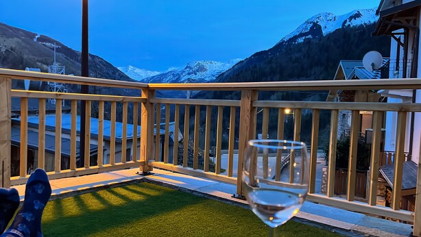 A balcony with a glass of wine, wooden railing, and a view of snow-capped mountains.