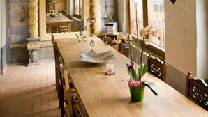 A dining area with a long wooden table, chairs, and a vase of flowers. The ceiling features colorful murals and a hanging lantern.