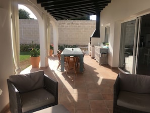 En overdækket terrasse med et bord og stole, et lille tekøkken og udsigt til haven.