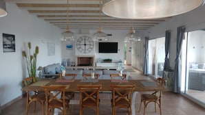 Un vaste espace dédié à la salle à manger, avec une longue table en bois et six chaises, une grande horloge murale et un téléviseur à écran plat fixé au-dessus d’une cheminée.