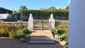 Un chemin de pierre mène à une entrée fermée par un portail en bois rustique, l’ensemble étant entouré d’un mur de pierre et d’une végétation luxuriante.