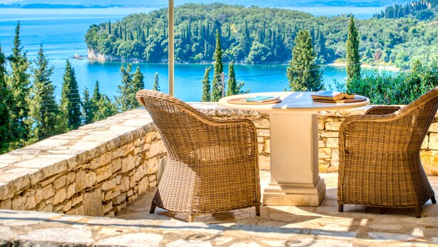 A terrace with wicker chairs and a table overlooking a lake and forested hills.