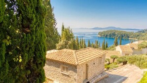 A stone building with a tiled roof, surrounded by trees and a view of a lake and mountains.