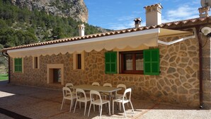 A stone building with a tiled roof, a dining area with white chairs, and a view of a mountainous landscape.