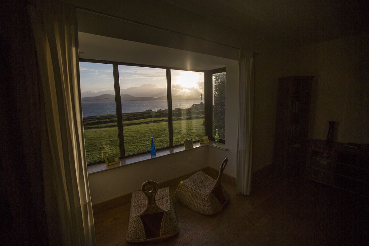 Una habitación con un ventanal grande que da a un paisaje cubierto de hierba y una masa de agua. Hay dos sillas de mimbre y una mesa pequeña con una botella azul en el alféizar.