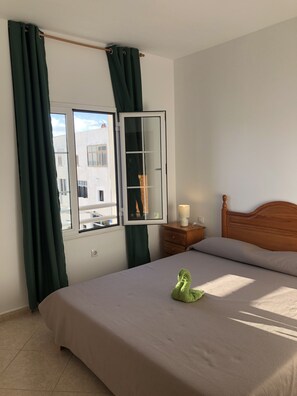 A bedroom with a bed, a wooden nightstand, a lamp, and a view through a window with curtains.