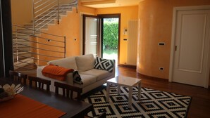 Uma sala de estar moderna com sofá, mesa de centro e tapete estampado. Uma escadaria é visível ao fundo, levando a uma área externa.
