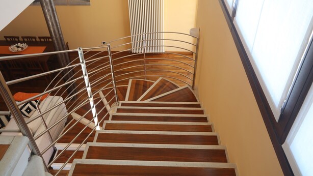 Uma escadaria em espiral com corrimãos de metal e degraus de madeira, que leva a um espaço com uma mesa de jantar e cadeiras.