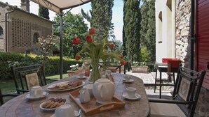 An outdoor dining area with a table set for breakfast, surrounded by greenery and a stone building.