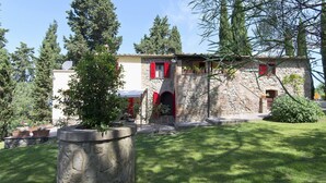 A stone building with red shutters, a small balcony, and a garden area with potted plants.