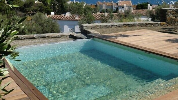 Piscina cristalina con bordes de madera, rodeada de edificios de estilo mediterráneo y vistas al mar.