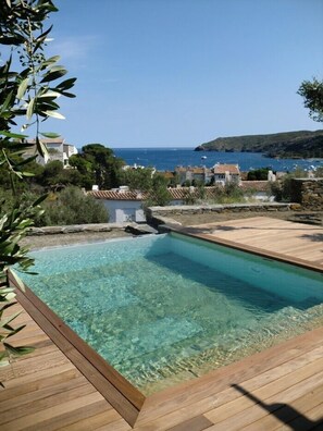 Een helder zwembad met houten randen, omringd door gebouwen in mediterrane stijl en uitzicht op zee.