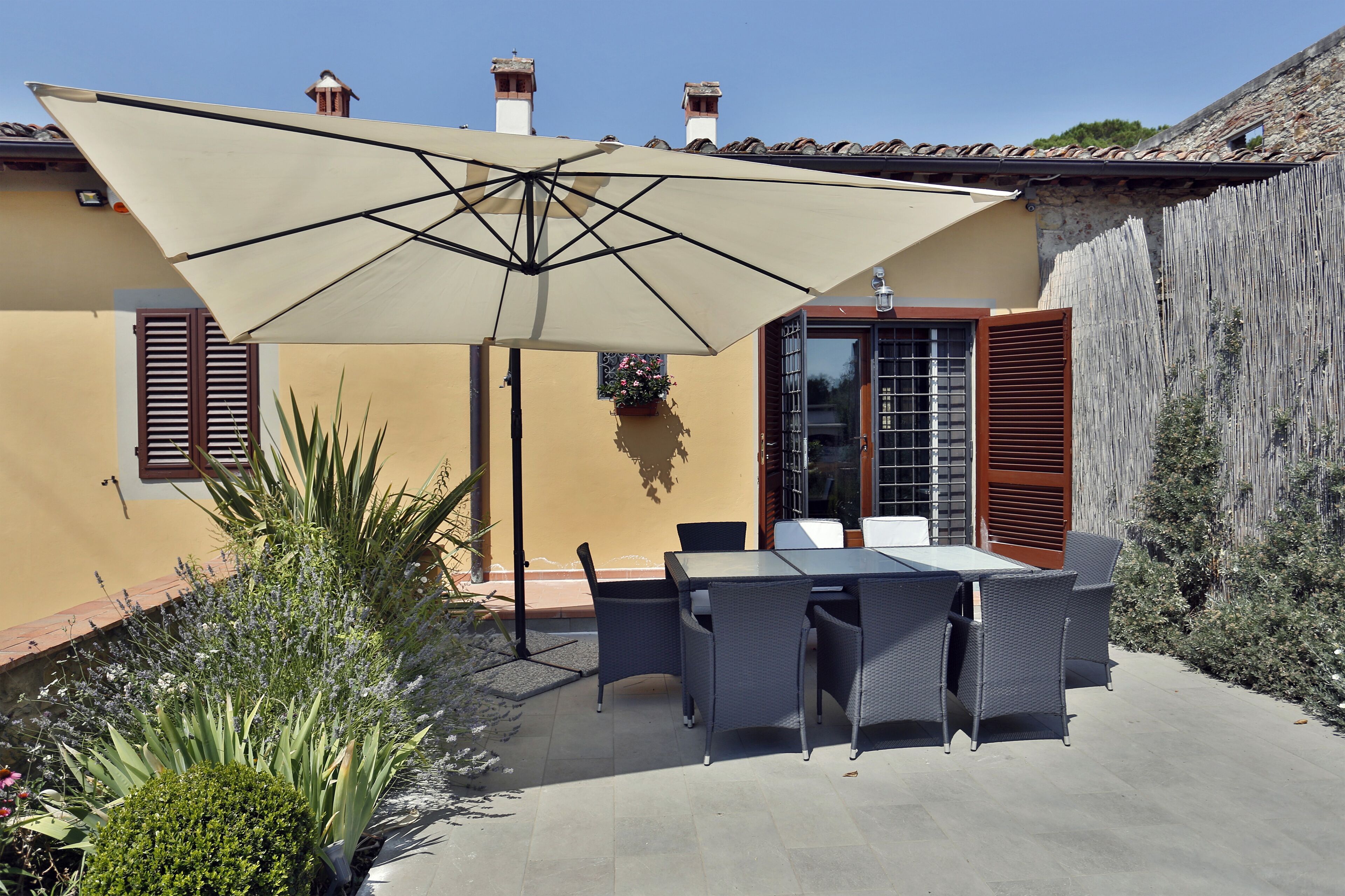 Una terrazza esterna con tavolo e sedie, un ombrellone grande e un edificio giallo con persiane e una finestra.