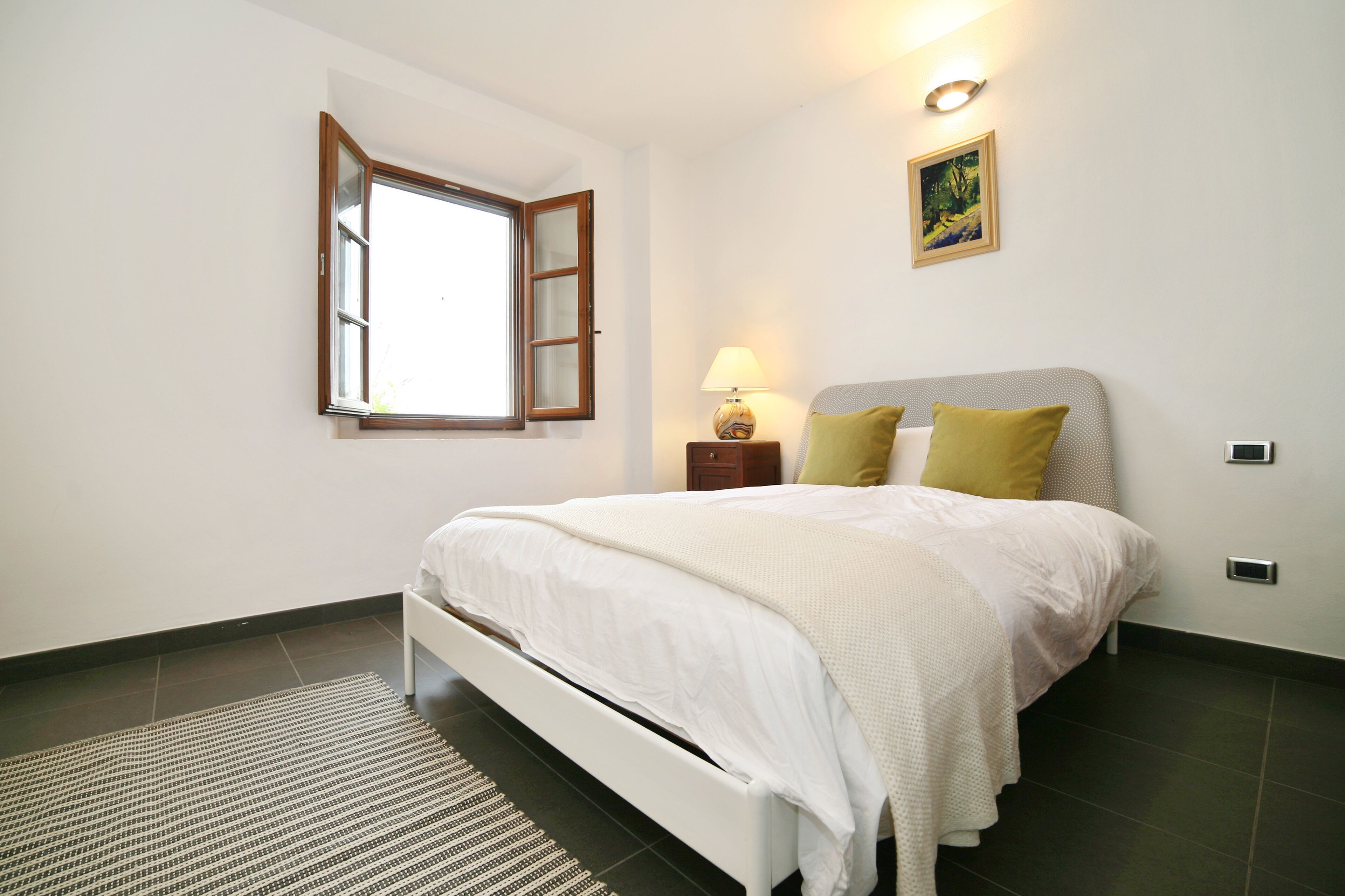 A bedroom with a bed, a nightstand, a lamp, a window, and a painting on the wall.