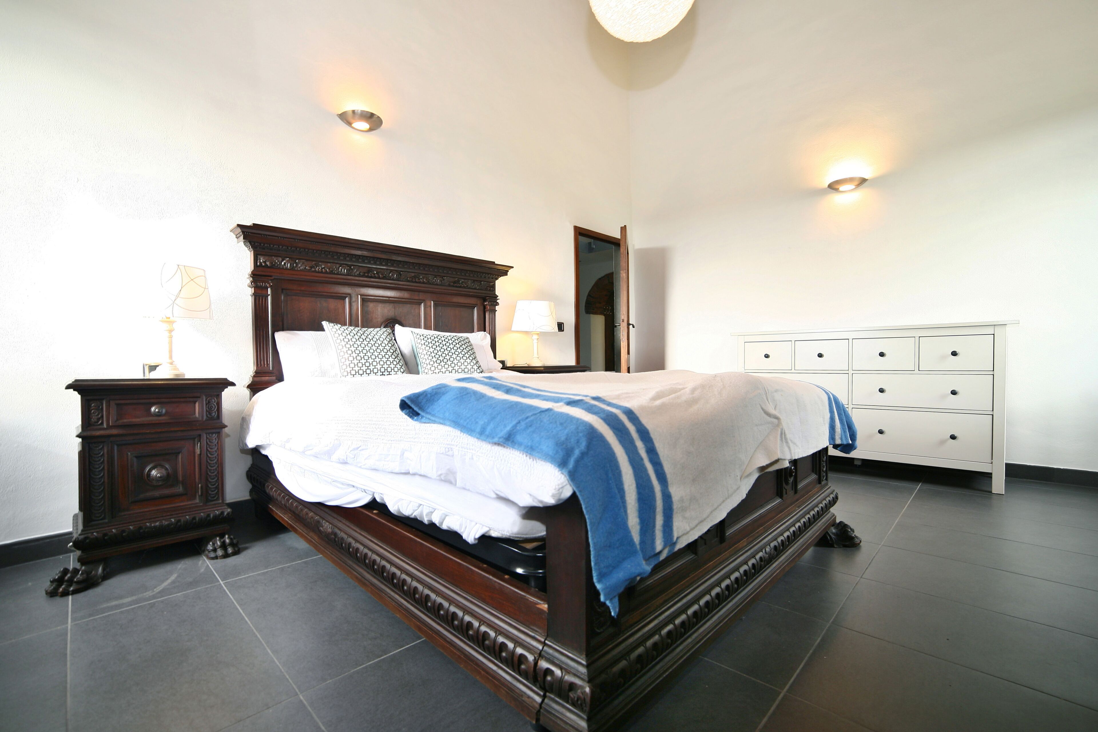 A bedroom with a large wooden bed, a nightstand, a dresser, and two wall-mounted lights.