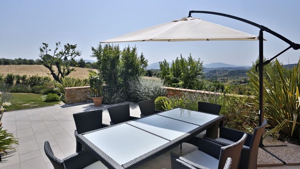 Um pátio externo com mesa e cadeiras, cercado por vegetação e vista para colinas ao longe.