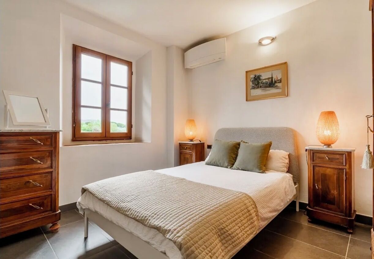 Une chambre à coucher comprenant un lit, des tables de chevet en bois, une commode, une fenêtre et un climatiseur fixé au mur.