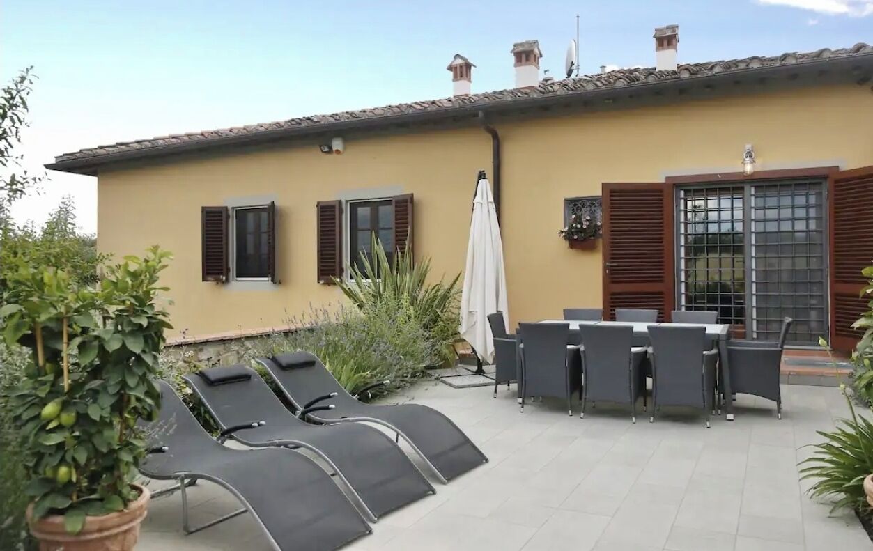 Ein Gebäude mit gelber Fassade, einer mit Fliesen belegten Terrasse mit Liegestühlen und einem Tisch sowie einem großen Fenster mit Rollläden.