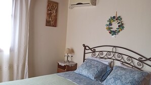 Een net opgemaakt bed met een groene sprei en blauwe kussens met een patroon, een houten hoofdbord, een wandairconditioner, een decoratieve krans en een klein nachtkastje met een lamp.