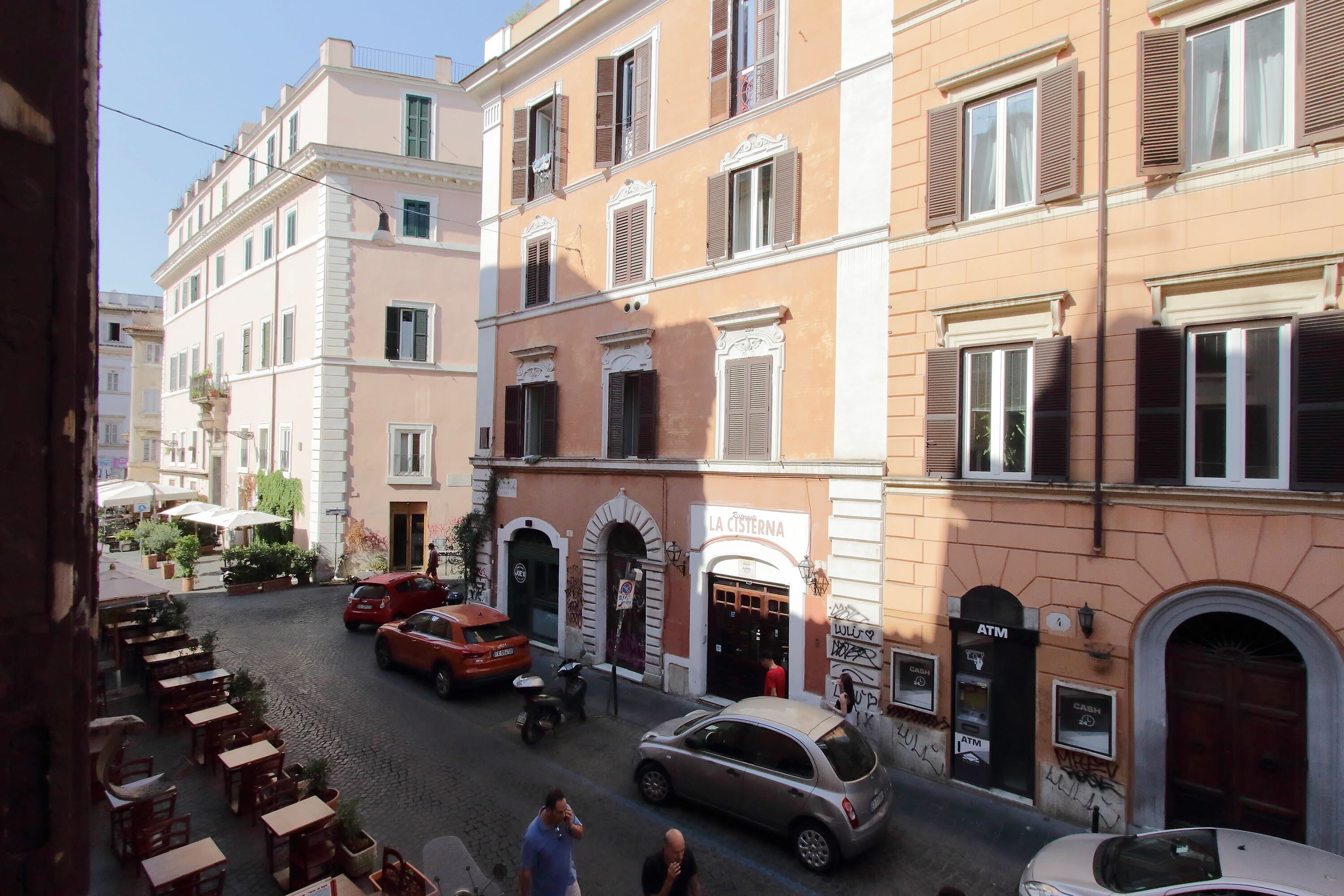 Una calle adoquinada flanqueada por edificios tradicionales, un café con mesas al aire libre y un automóvil estacionado frente a un edificio con cajero automático.