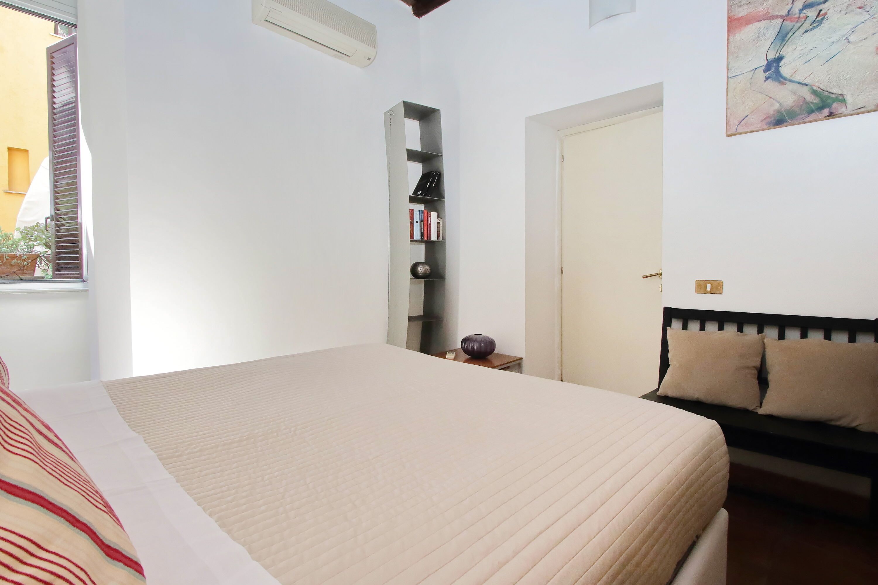 Un dormitorio con una cama grande, una estantería, una mesita con un florero y un sofá con cojines.