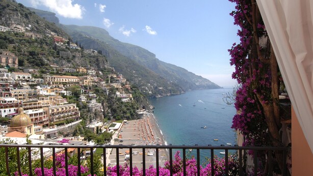 A coastal town with buildings on a hillside, a beach with umbrellas, and boats on the sea.