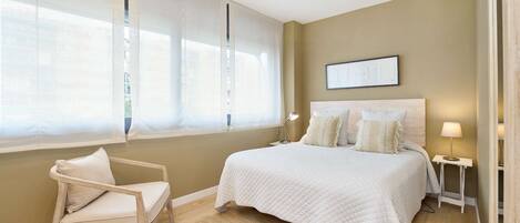 A bedroom with a bed, a chair, a bedside table, and a window with sheer curtains.
