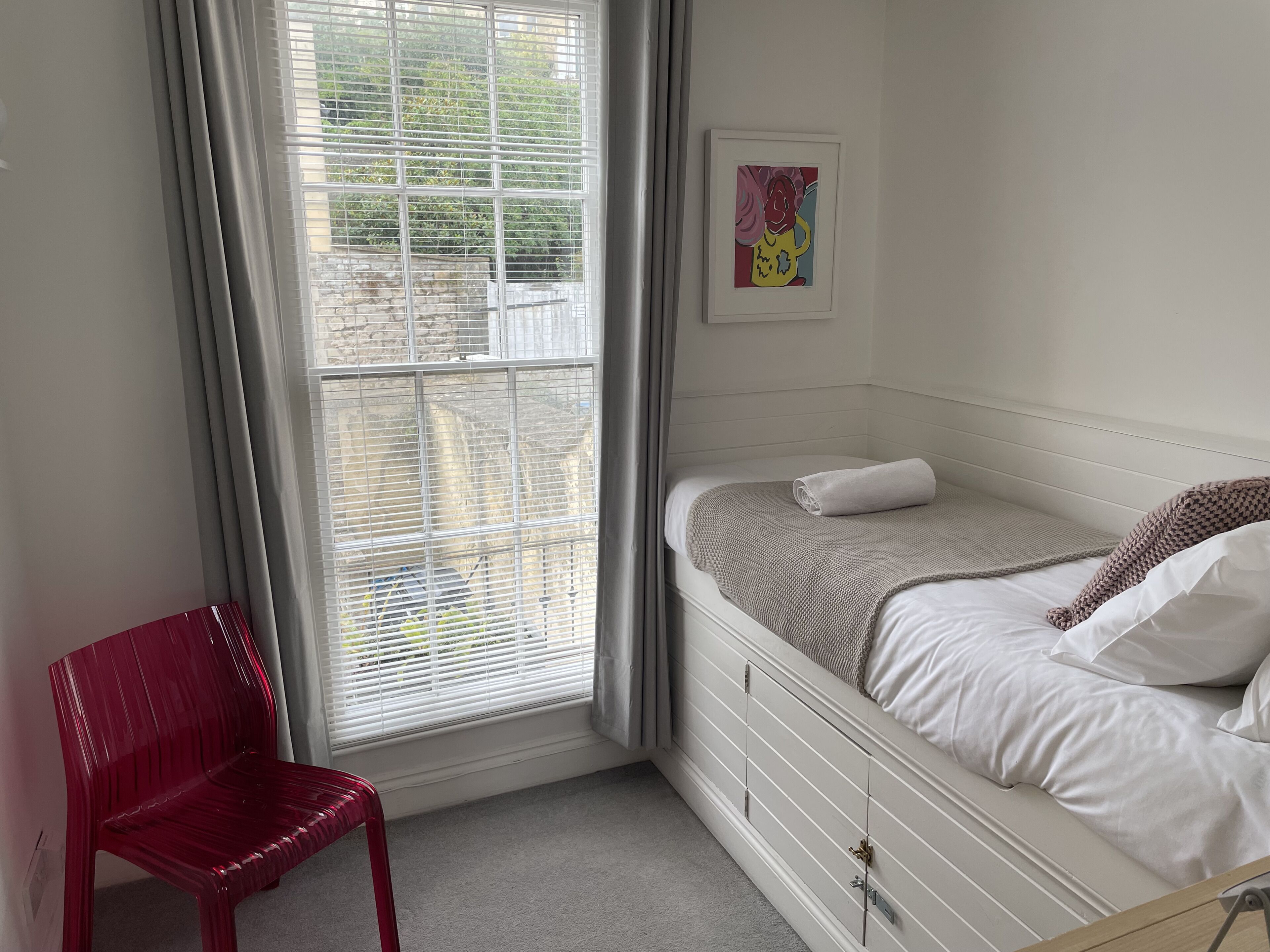 Een slaapkamer met een bed, een raam met zonwering, een rode stoel en een ingelijste foto aan de muur.