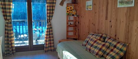 Een slaapkamer met een bed, ruitersgordijnen, een houten wand en een raam met uitzicht op tuinmeubilair.