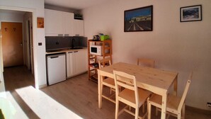 A compact kitchen with white cabinets, a microwave, and a sink. There is a wooden table with chairs, a small shelf with a kettle, and two framed pictures on the wall. - Studio - Nevache (Névache)