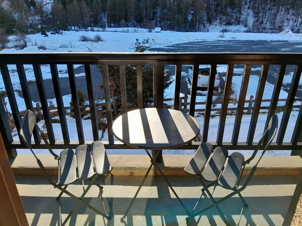 A balcony with a table and four chairs, overlooking a snowy landscape with a river and trees.