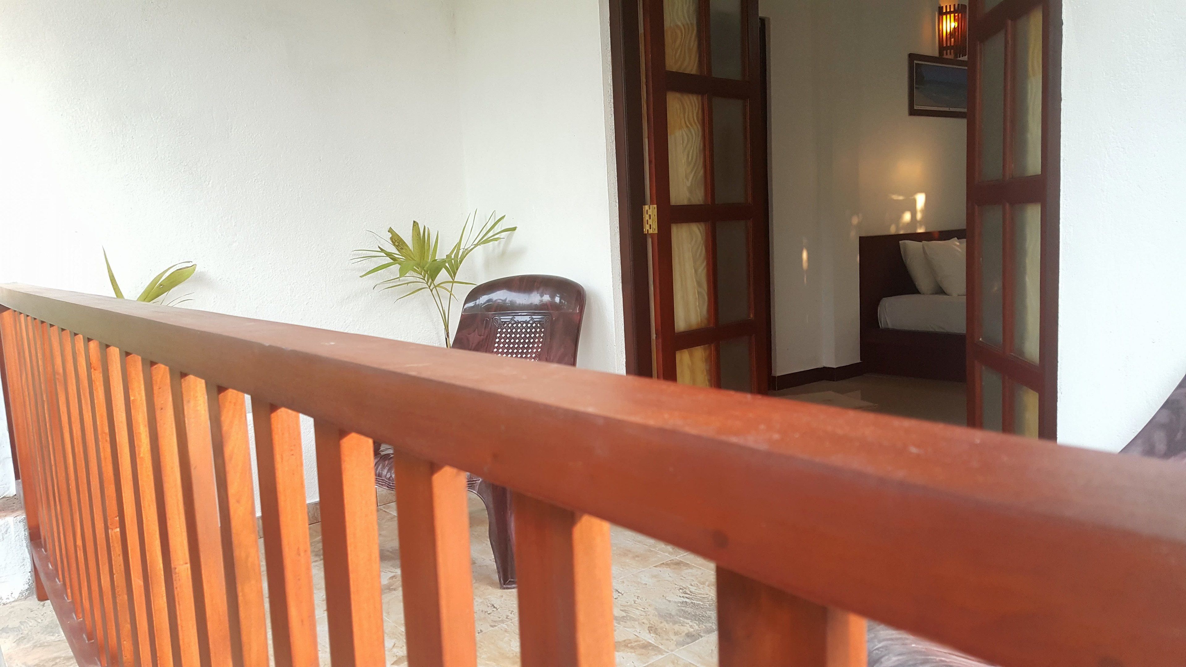 A wooden balcony railing, a potted plant, a chair, and a glimpse of a room with a bed and a nightstand.