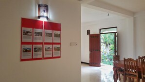 A hallway with a bulletin board displaying multiple informational posters, a wooden door leading outside, and a seating area with wooden chairs. - Villa Happy Home (Bentota)