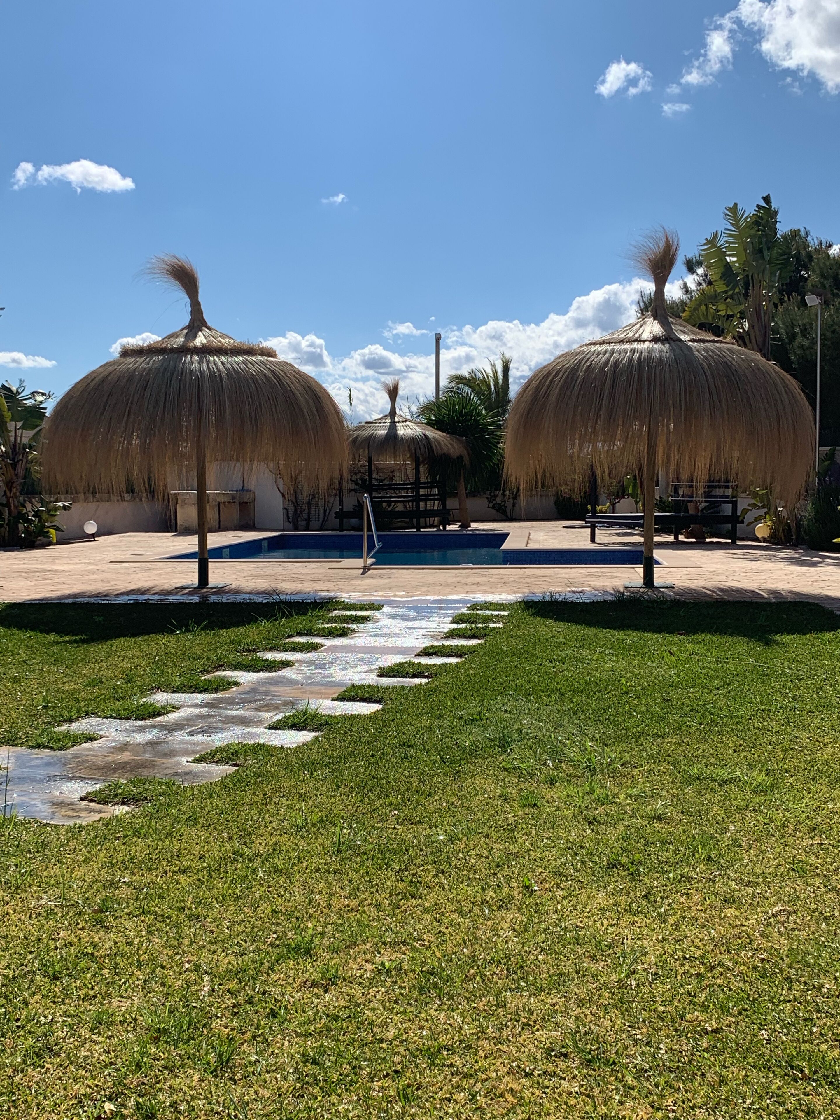 Un espace piscine agrémenté de parasols en chaume, un ciel d’un bleu limpide et un chemin bordé de pelouse.