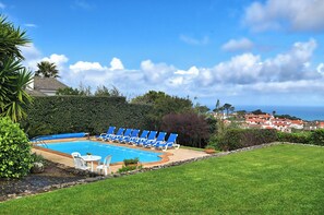 A swimming pool with blue lounge chairs and a white table, surrounded by greenery and a view of houses in the distance.