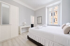 Un dormitorio con cama, escritorio, espejo y ventana con vista a edificios.