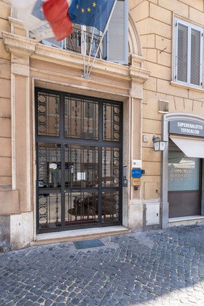 Fachada de edifício com um grande portão metálico ornamentado, uma placa com a inscrição 'Superenalotto Totocalcio' e uma rua de paralelepípedos.