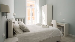 Un dormitorio con cama, mesitas de noche, escritorio y ventana con vista a un edificio.