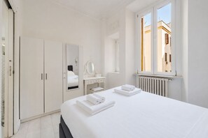 Une chambre d’hôtel comprenant un lit, une coiffeuse avec un miroir, une armoire et une fenêtre donnant sur des immeubles.