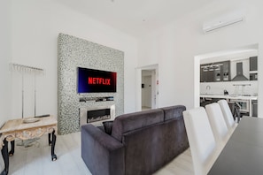 Un moderno salón con una televisión de pantalla plana montada en una pared de mosaicos, un sofá de color oscuro y una mesita de madera con un control remoto.