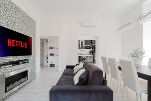 Un salon moderne avec un téléviseur à écran plat, un canapé foncé, un coin repas avec des chaises blanches et une cuisine en arrière-plan.